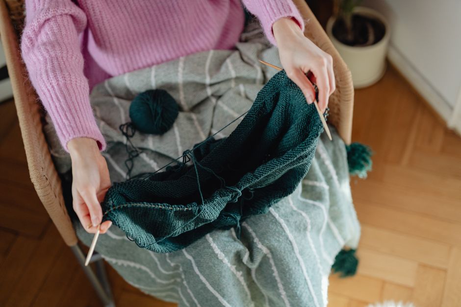A young woman knitting a green wool garment in a comfy armchair indoors.