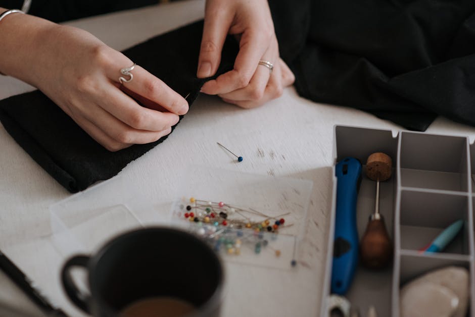 Unrecognizable female dressmaker using pins on black textile while sewing at table with container of professional instruments in modern studio