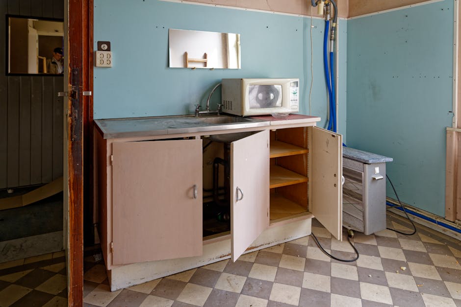 An abandoned kitchen with open cabinets and a microwave on a worn countertop.