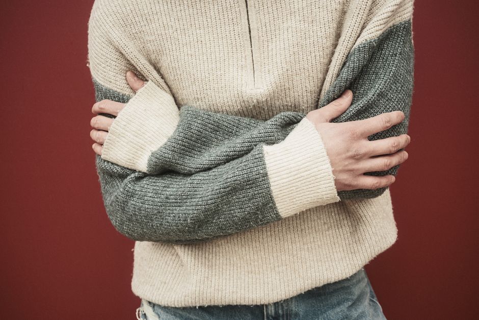 Close-up of a person in a cozy sweater with arms crossed, standing against a red wall.
