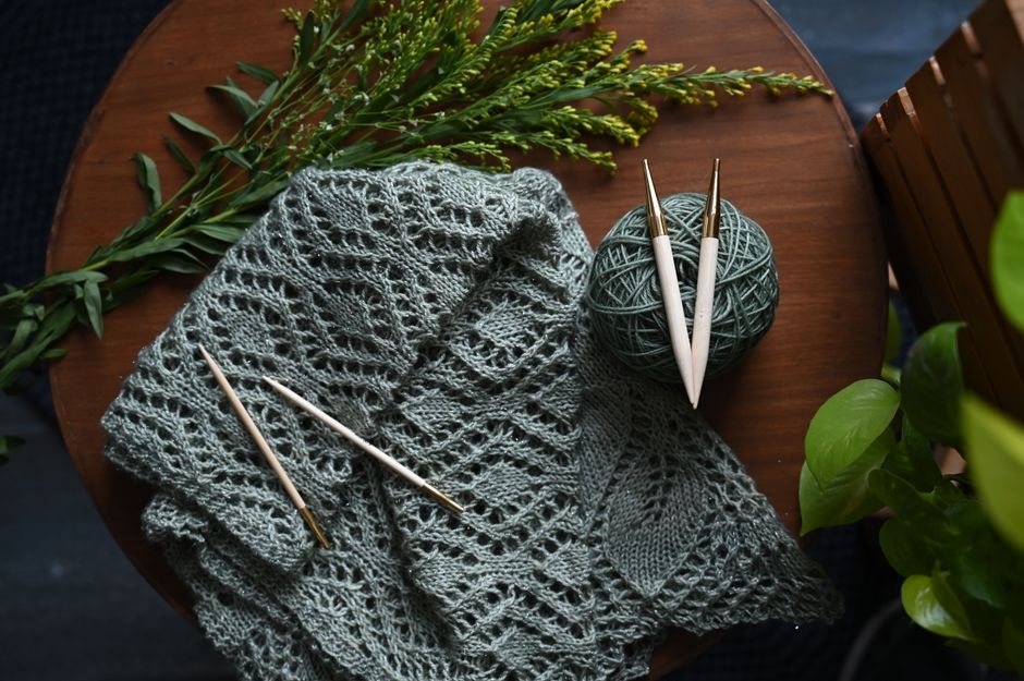 Green knitted shawl with crochet hooks on a wooden table, surrounded by greenery.