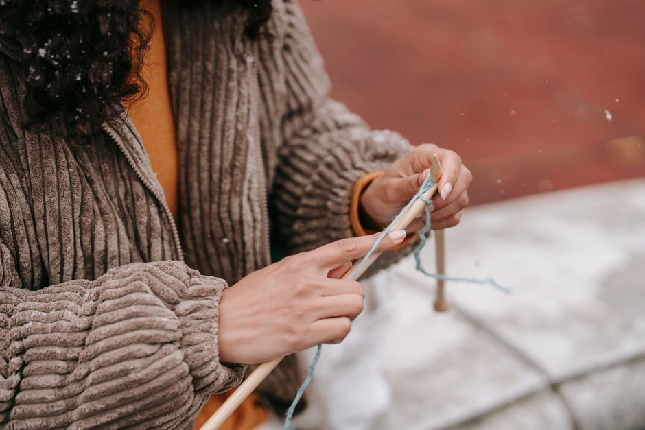 Crop anonymous female with dark curly hair knitting with thread of woolen yarn and needles wearing corduroy light brown jacket and orange t shirt